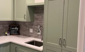 Custom laundry cabinets in Algonquin Highlands, ON, with a tall sage green shaker cabinet beside a stainless undermount sink, upper cabinets with crown moulding, a white countertop and grey brick-look backsplash, by Palmer's Woodworking.