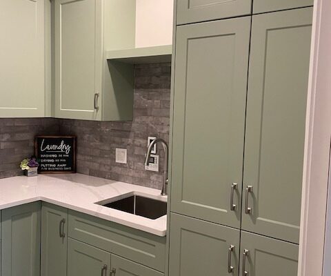 Custom laundry cabinets in Algonquin Highlands, ON, with a tall sage green shaker cabinet beside a stainless undermount sink, upper cabinets with crown moulding, a white countertop and grey brick-look backsplash, by Palmer's Woodworking.
