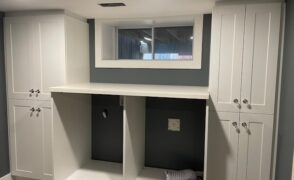 Custom white shaker laundry room cabinets with tall double-door cabinets on each side, a long white counter over the washer and dryer openings and a white-trimmed basement window, designed, built and installed by Palmer's Woodworking in Guelph, Ontario.