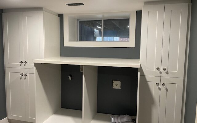 Custom white shaker laundry room cabinets with tall double-door cabinets on each side, a long white counter over the washer and dryer openings and a white-trimmed basement window, designed, built and installed by Palmer's Woodworking in Guelph, Ontario.